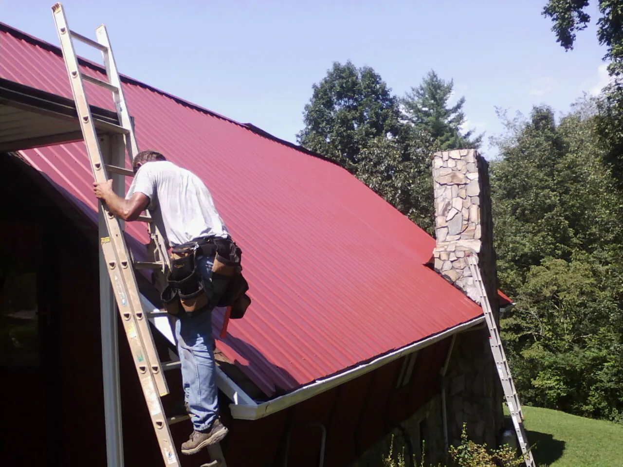 Roofer on a ladder beside a red metal roof and stone chimney — Soffit & Fascia Repair by Roofing Brothers in Fraser